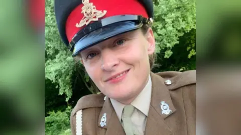 Handout A woman in smart military dress smiles at the camera. She is leaning slightly to one side and has a half-smiling expression. She wears a brown jacket with silver detailing and a black hat with a red band of colour above the visor. She is in front of greenery.
