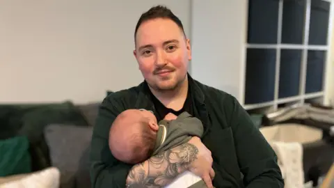 BBC/Olivia Richwald A parent with short dark hair and a goatee beard holds a young baby.
