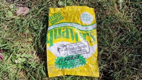 An old quavers crisp packet in yellow, green and white colours, on a patch of green grass