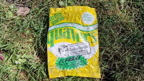 Cenk Albayrak-Touye An old quavers crisp packet in yellow, green and white colours, on a patch of green grass