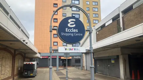 BBC The entrance to Eccles Shopping Centre with a large sign in the precinct. In the background and to the right hand side are shuttered up shops. There is one lone person in the area, otherwise it is empty.