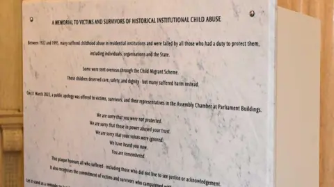 Pacemaker Memorial plaque at Stormont acknowledging the suffering caused to thousands of people who were abused in children's homes and other residential institutions. The plaque is white and has black writing etched on it.