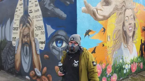 A colourful mural on two sides of a wall depicts a dark and lighter side. On the right is a woman with blonde hair smiling, blue skies, sun, flowers and birds behind her. On the left is a man holding his head in his hands with words including 'cancer, stress, no, health' written in a lung-diagram on a dark blue background. A man is stood in-between the two walls in a green coat, grey meanie, mask and holding a paint can.