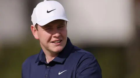 Getty Images Head and shoulders shot of a smiling Robert MacIntyre. He is wearing a dark blue polo shirt and white baseball cap. 