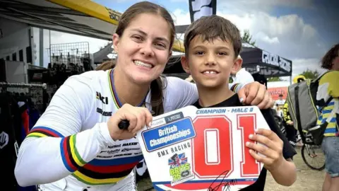 Archie Fisher is on the right of picture, posing with Team GB rider Beth Shriever. Beth is wearing a white racing suit. They are holding a sign saying 'British Championships BMX Racing Champion Male 8', a title Archie won at the British BMX Championships in Kent in 2025.