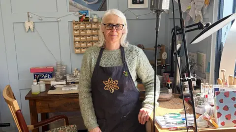 LDRS Claire Howard is wearing a blue apron with a yellow flower pattern on it. The apron is over a green jumper. She's also wearing glasses and we can see she is in an arts workshop of some kind.