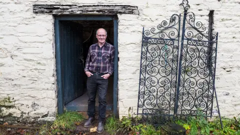 Mark Lewis Paul Dennis stands in the doorway of a whitewashed stone outbuilding. The wooden blue door is open behind him and there is a faded black lintel across the top. To his right, leaning against the wall, is a pair of wrought iron gates with lots of curling interlinked spirals. There are grass and weeds growing at the edge of the building.