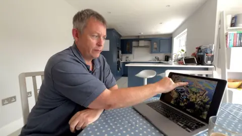 A man with grey hair and wearing a grey T-shirt sitting at a kitchen table and pointing to a photo on his laptop screen. It's an aerial photo showing a flooded forested area.