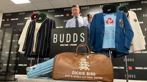A person with short brown hair, blue shirt and tie stands behind a black podium branded “BUDDS” in a showroom or auction-style setting. On either side of the podium are display mannequins wearing cricket memorabilia, including blazers, shirts, and caps. In the foreground, a large brown leather cricket bag marked “DICKIE BIRD – TEST & WORLD CUP FINAL UMPIRE” sits on top of a silver equipment case, with light-blue cricket stumps leaning beside it. The backdrop is covered with repeating “BUDDS.COM” and “BUDDS” logos.
