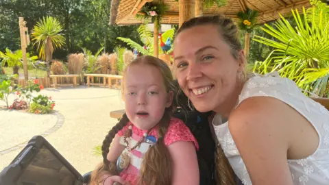 A mother and daughter sitting in the garden.
The six-year-old girl is sitting on the left and has two plaits in her hair. The mum is on the right. She has her hair in a ponytail and is wearing a white top. 