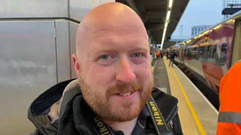 A man with an auburn beard and mustache. He wears a gray coat and has a camera strap around his neck. He is standing on a train platform.