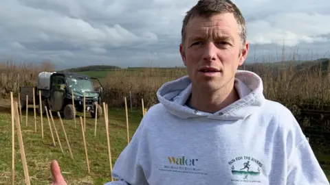The picture shows a person standing in a field that appears to be part of a tree‑planting or conservation project. The ground behind them is lined with rows of wooden stakes, likely marking spots for young trees or protective tubes. A utility vehicle with equipment on the back is parked further up the field. The person is wearing a light grey hoodie with environmental and charity‑themed logos on the front, and they are gesturing with their hands as if explaining or discussing the work being done. The landscape around them is open countryside under a cloudy sky.