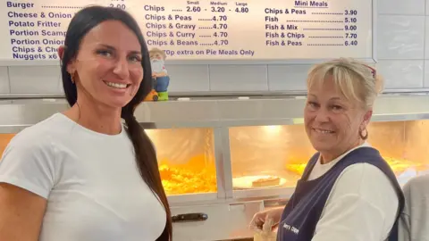 Two women stand beside a chip shop fryer. They are dressed in white tops and the woman on the right has a navy tabards. There is a blue and white illuminated price list above the fryer on a white tiled wall.