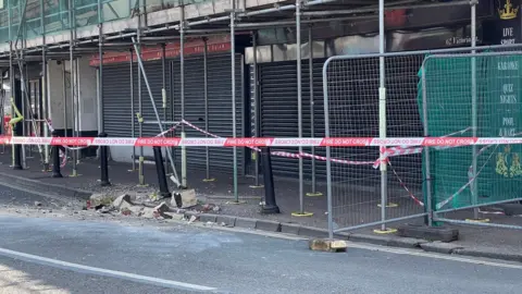 Debris on the road and pavement behind 'Fire do not cross' red and white tapewith scaffolding and fencing visible. 