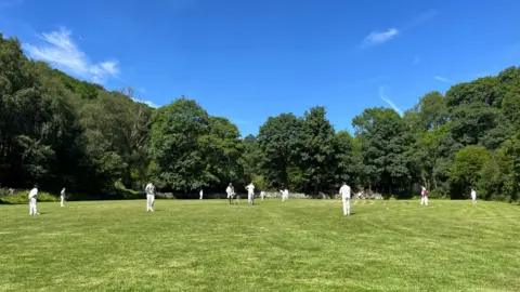 Hebden Bridge Cricket Club A picture of Hebden Bridge Cricket Club paying a game. The players are wearing cricket whites and they are surrounded by trees.