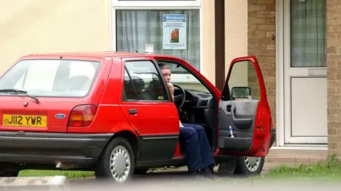 Medios de comunicación de PA: Un hombre medio sentado en un Fiesta rojo. Lleva vaqueros azul oscuro. Está sentado de lado en el asiento del conductor. La puerta del conductor está abierta y el hombre tiene los pies en el suelo.
