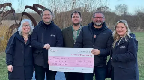 A group of people, including Emily Routledge, who has blonde hair and is wearing a dark coat, are standing outside and holding a large cheque for £326,543.00.