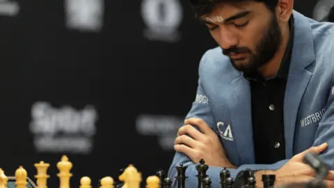 EPA Gukesh Dommaraju gazes down at the chess board at the game on Thursday. He is wearing a blue jacket and a black shirt and has his arms folded, with his elbows resting on the table