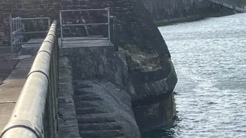 The stairs, which lead down to the sea and are heavily corroded down the side closest to the water, leaving them smooth. They are fenced of by steel fencing with large gaps between the bars. There is, as reported, no signage visible in the photo.