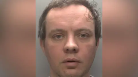 Police mugshot of Rick Parker - a man with wavy dark hair. He is clean shaven and is looking directly at the camera.