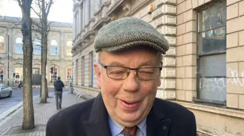 John Gray, wearing a cap, glasses, dark jacket, blue shirt and brown tie