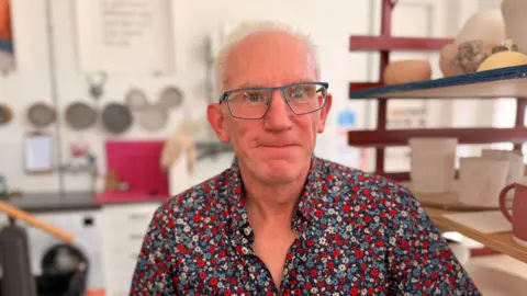 A man with grey hair and glasses, he is wearing a colourful flowered shirt and is looking at the camera smiling.