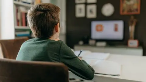 The back of a child sitting at a desk looking at a computer screen. Has brown hair, wearing a long sleeve green top and writing in a booklet.