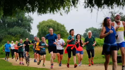 A group of people running as part of a parkrun. There are men and women of all different ages. One is running with a dog. They are all wearing running clothes - shorts, vests and trainers.