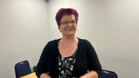 Jessica Bayley/ BBC A woman with short dyed red hair and glasses stands looking towards the camera. She has a slight smile and is wearing a black top with white leaves on it, with a black cardigan over.
