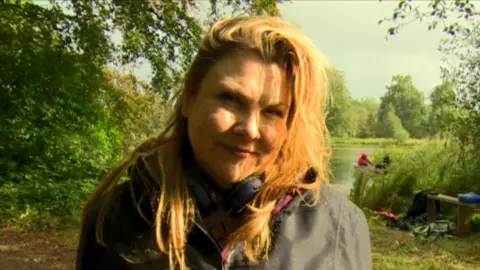 A woman with mid length hair smiles at the camera. She is wearing a waterproof jacket and behind her in the distance is a lake and green shrubbery. A golden light from the sun is hitting her on the right side of her face.