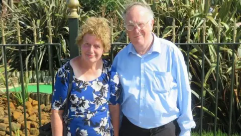 Family handout Lois and John McCullough, who are standing in front of a mini golf course. They are both dressed in blue and smiling at the camera.