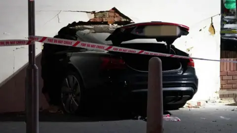 A black car halfway in a property which is painted white. Its boot is up and it is behind red and white tape which says fire do not cross.