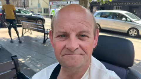 Christopher Smalley, 54, has a mainly bald head, with some dark hair on the sides of his head. He is sitting in a mobility scooter and behind him is a town square, with benches and cars passing on the nearby roadway.