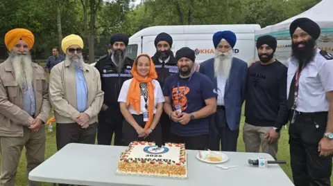 West Midlands Police A group of eight men and one woman stand by a table in an outdoor area. There is a cake on the table. Some of the people are wearing police uniforms. 