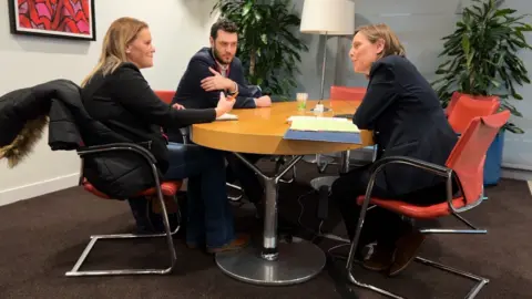 Micala sits at a large round table in an office talking with Joe Morris and Jess Phillips. Micala has long blond hair, Joe a neat beard and short black hair and Jess shoulder length brown hair.
