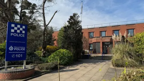A red brick building with POLICE above the entrance. A path leading up to the front is lined with trees and bushes. A blue sign at the near end of the path indicates it belongs to Warwickshire Police.