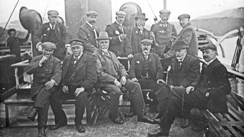 A black and white photograph showing a group of smartly dressed men wearing hats on the deck of a paddle steamer sailing on Loch Ness. Some of them are smoking cigarettes. 