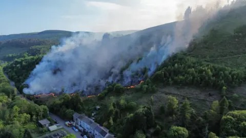 Gavin Mathews An aerial image shows a large area of grass on a hillside burning, with a line of orange flames towards the bottom of the hill as smoke billows into the air. A row of terraced houses can be seen close to the flames, with a road running along the bottom of the hill. 