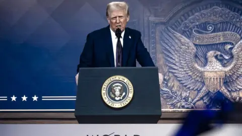 President Donald Trump speaking via video link to World Economic Forum. He stands before a blue background and stand before a podium with a presidential seal  