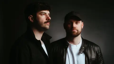 Glasgow 2026 Two men with dark hair, beards and moustaches wearing a white t-shirts and black jackets. One is facing forward and looking at the camera while the other is looking to the right away from the camera.