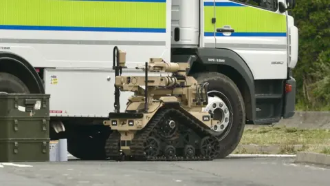 A small beige-coloured robot is stationery on the road. It is next to a bomb disposal vehicle. 