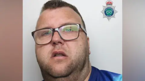 Satffordshire Police Custody image of Dale Cole a man with glasses and stubble on his chin and neck. He has short dark brown hair and is wearing a blue tie-dye tshirt as he stands against a grey background