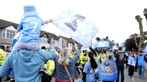 PA Media A man wearing a blue hoodie holds a small boy on his shoulders wearing a Coventry City kit and waving a flag that reads "Coventry champions". The pair are surrounded by other Coventry City fans as the bus drives past.