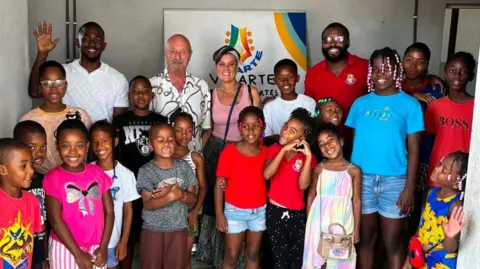 Chris Backhouse Backhouse stands alongside adults and children in Cape Verde. They smile and wave at the camera.