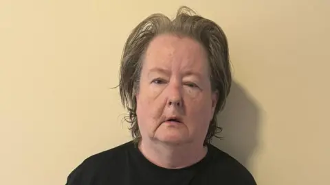 Handout A close-up of a woman with short dark grey hair looking at the camera. She stands in front of a white wall and wears a black t-shirt.