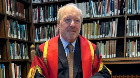 BBC Sir Alan Bates sits in Bangor University library, wearing red, gold and black doctorate gown and a black suit, with a blue shirt and brown tie. Behind him are shelves of books.