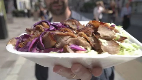 A man who is out of focus holding a kebab with brown meat, green lettuce and red (purple) cabbage. The packaging is white.