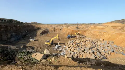 BBC A quarry site with huge piles of rock and a couple of yellow earthmovers. The brown earth contrasts with the blue sky