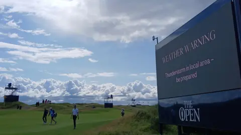 BBC Weather Watcher A large sign at a green at The Open saying "Weather Warning. Thunderstorm in local area - be prepared". Sun is shining with clouds approaching. People are walking along the green.
