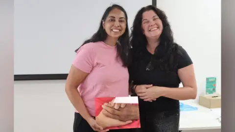 Supplied Laila Fazluddin and Rachel Marsh are standing side-by-side and both smiling brightly at the camera. Fazluddin, who has long brown hair and is wearing a pink top, is holding a picture of two hands embracing. Marsh is wearing a black t-shirt and also had long brown hair.